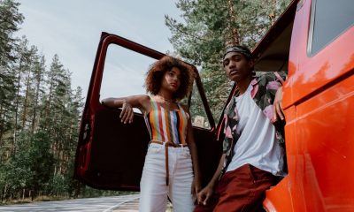 Two people lean against a red pickup truck on the side of a rural highway lined with trees.