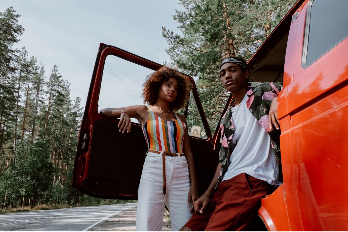 Two people lean against a red pickup truck on the side of a rural highway lined with trees.