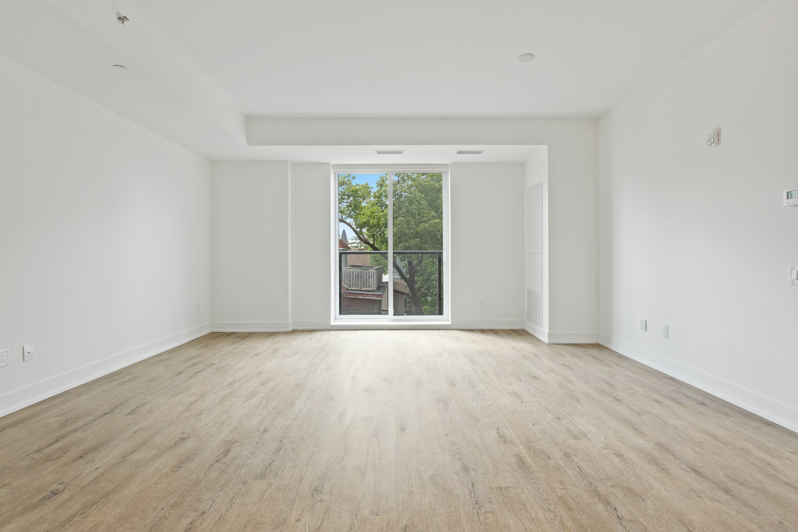 An empty unfurnished condominium unit with pale wood flooring, white walls and a balcony door