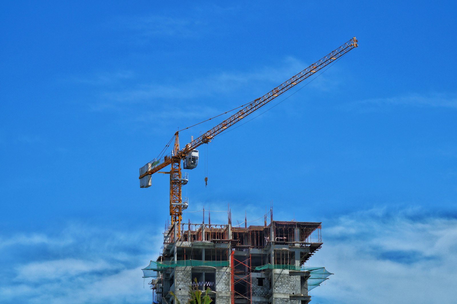 A tower crane rises above the upper floors of a concrete highrise under construction against a blue sky