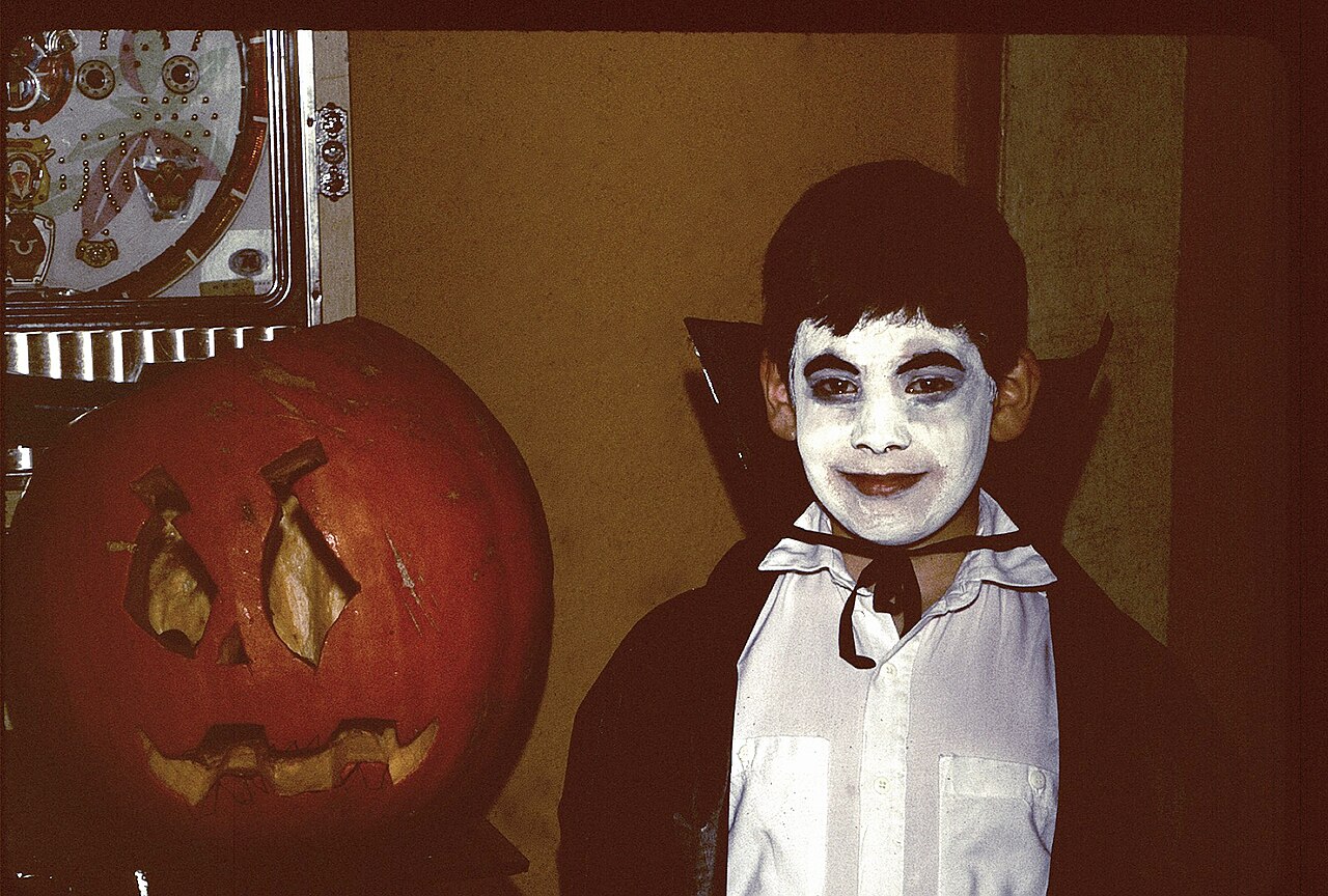 Child in a vampire costume with cape and white face paint beside a carved pumpkin