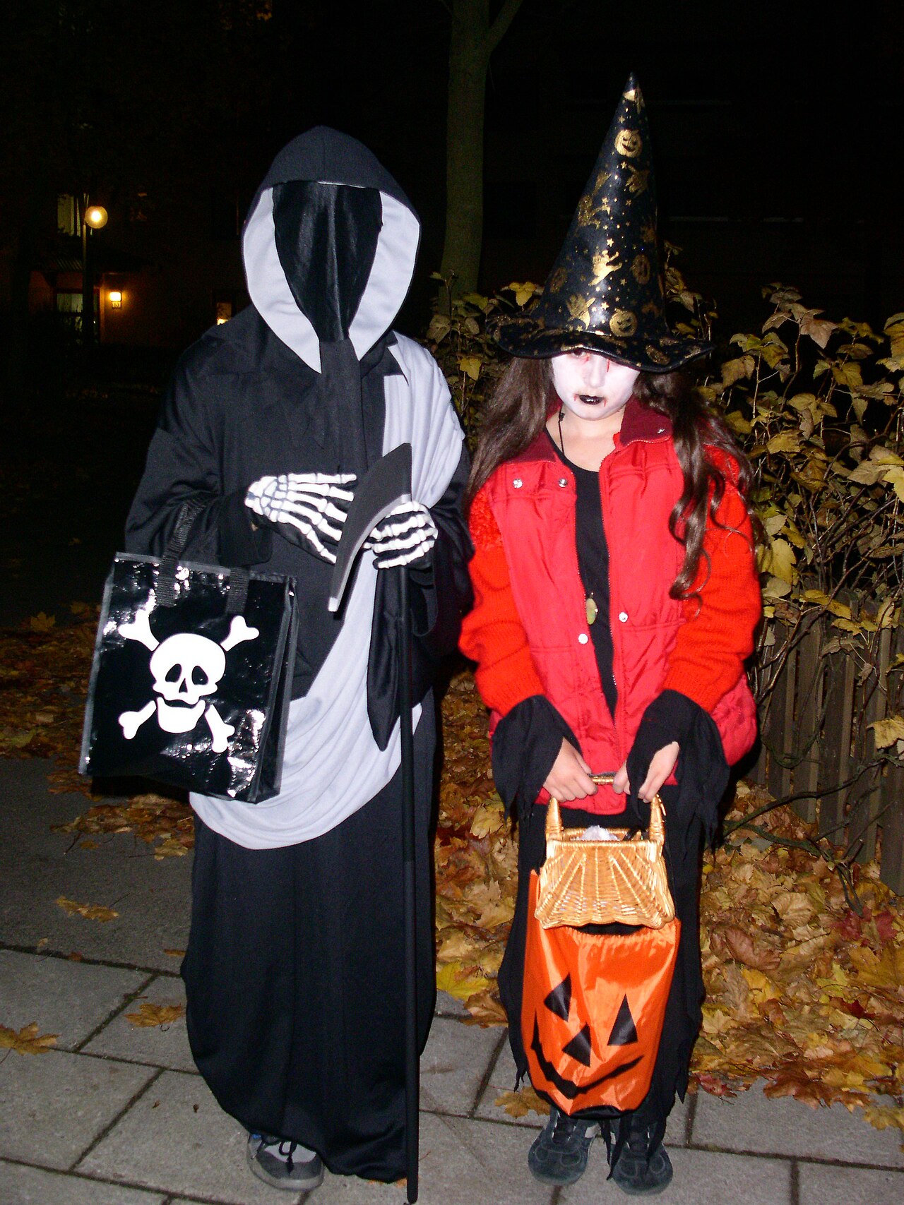 Two children in a skeleton costume and a witch costume holding candy bags at night