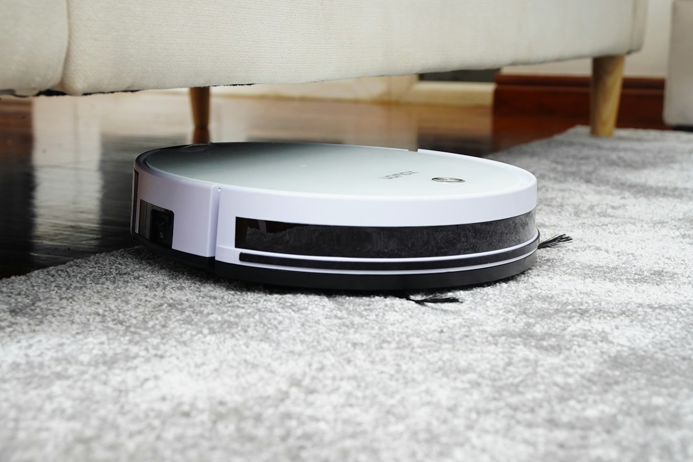 A white robot vacuum cleaning a grey carpet beside a hardwood floor