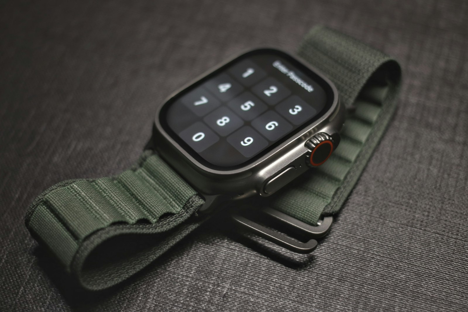 Apple Watch Ultra with a green Alpine Loop band and orange Action button resting on a dark surface