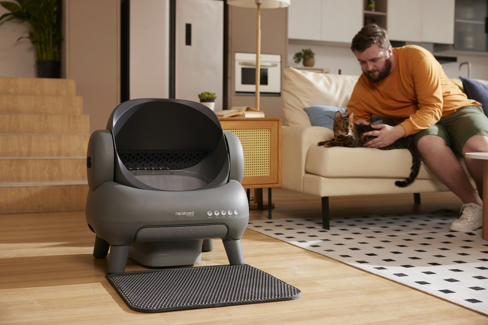 Man on a couch petting a Bengal cat beside a grey Neakasa automatic litter box