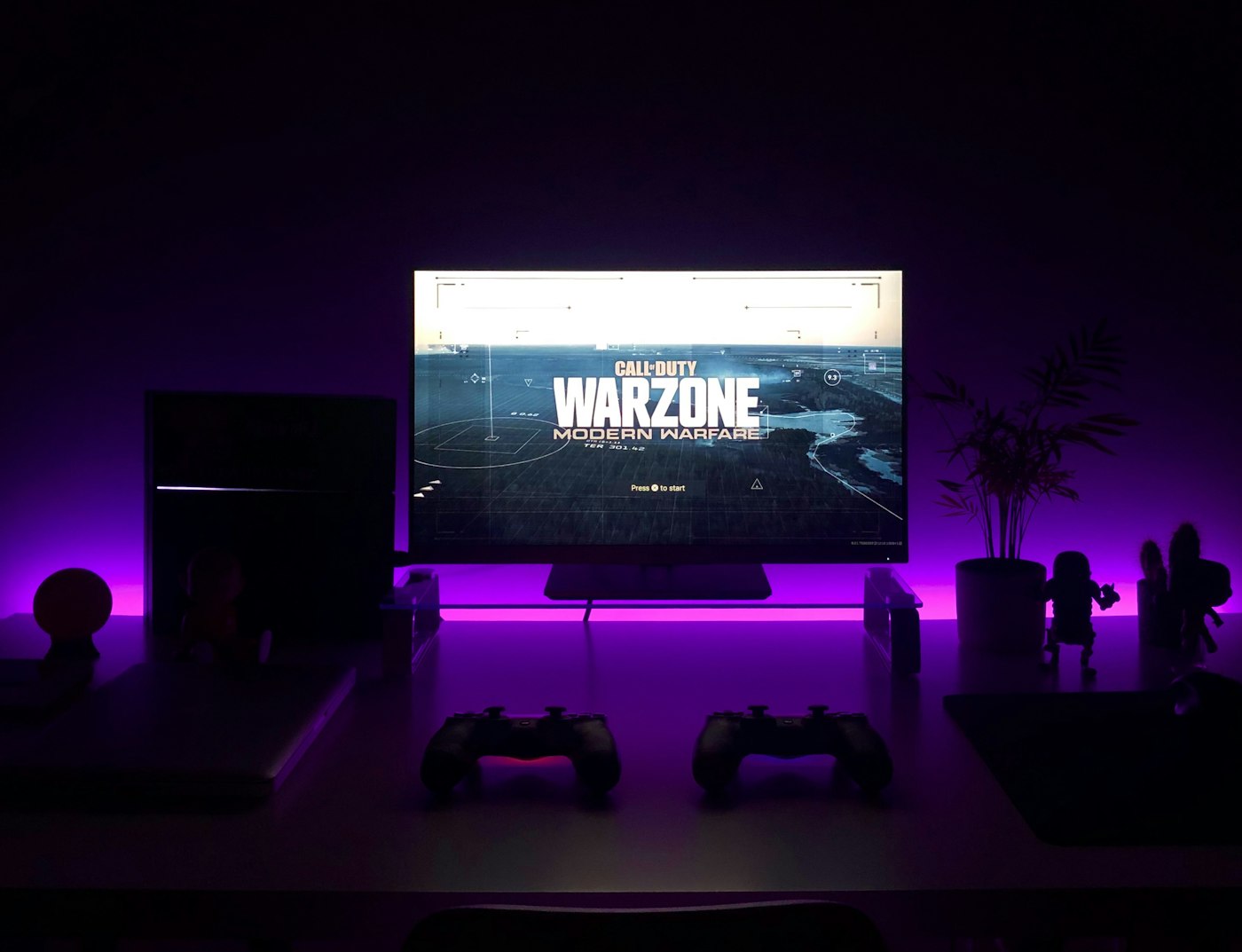 Two game controllers on a desk in front of a TV with purple LED lighting