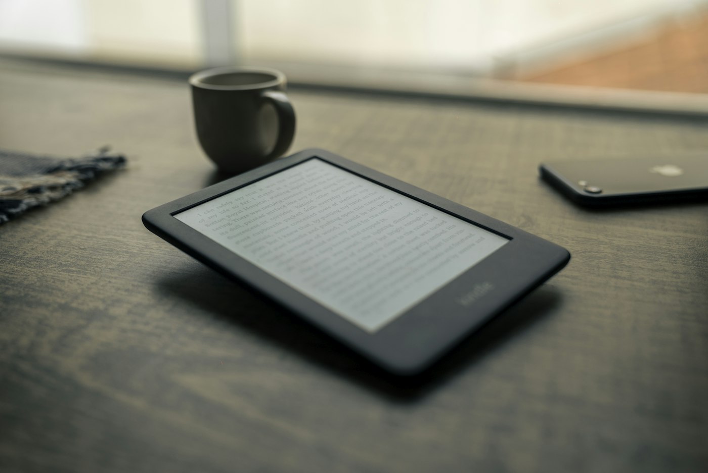 Kindle e-reader lying on a wooden table beside a mug and a phone