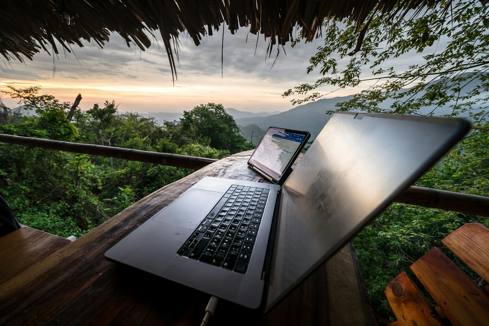 Laptop and phone on a wooden table overlooking a forested hillside at sunset