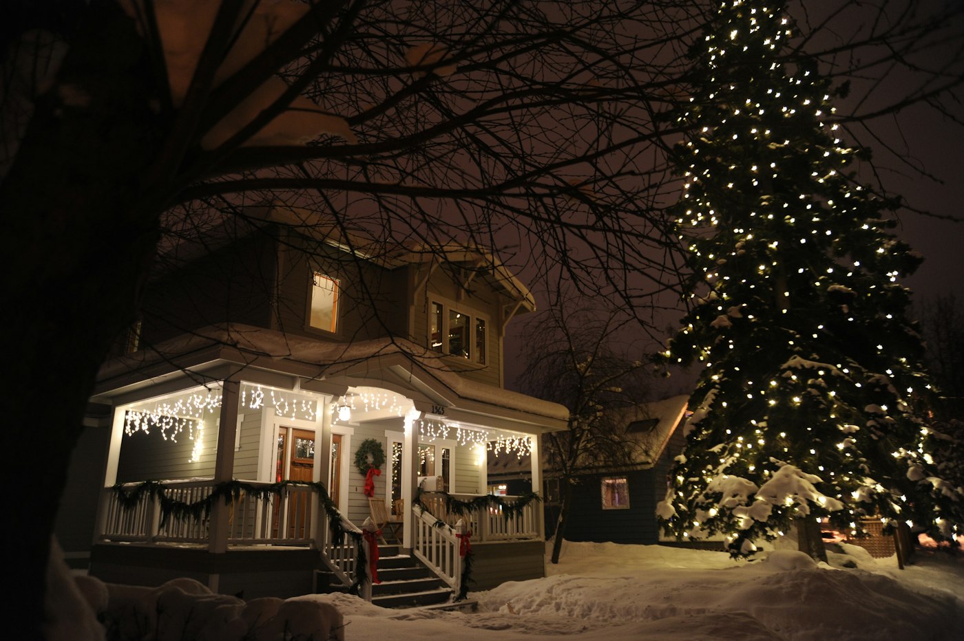 Spruce tree lit with white Christmas lights beside a house under fresh snow