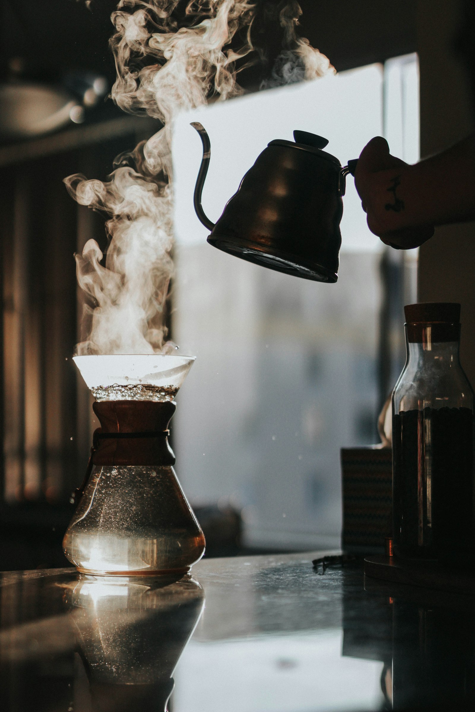 Hand pouring hot water from a gooseneck kettle into a steaming pour-over coffee maker