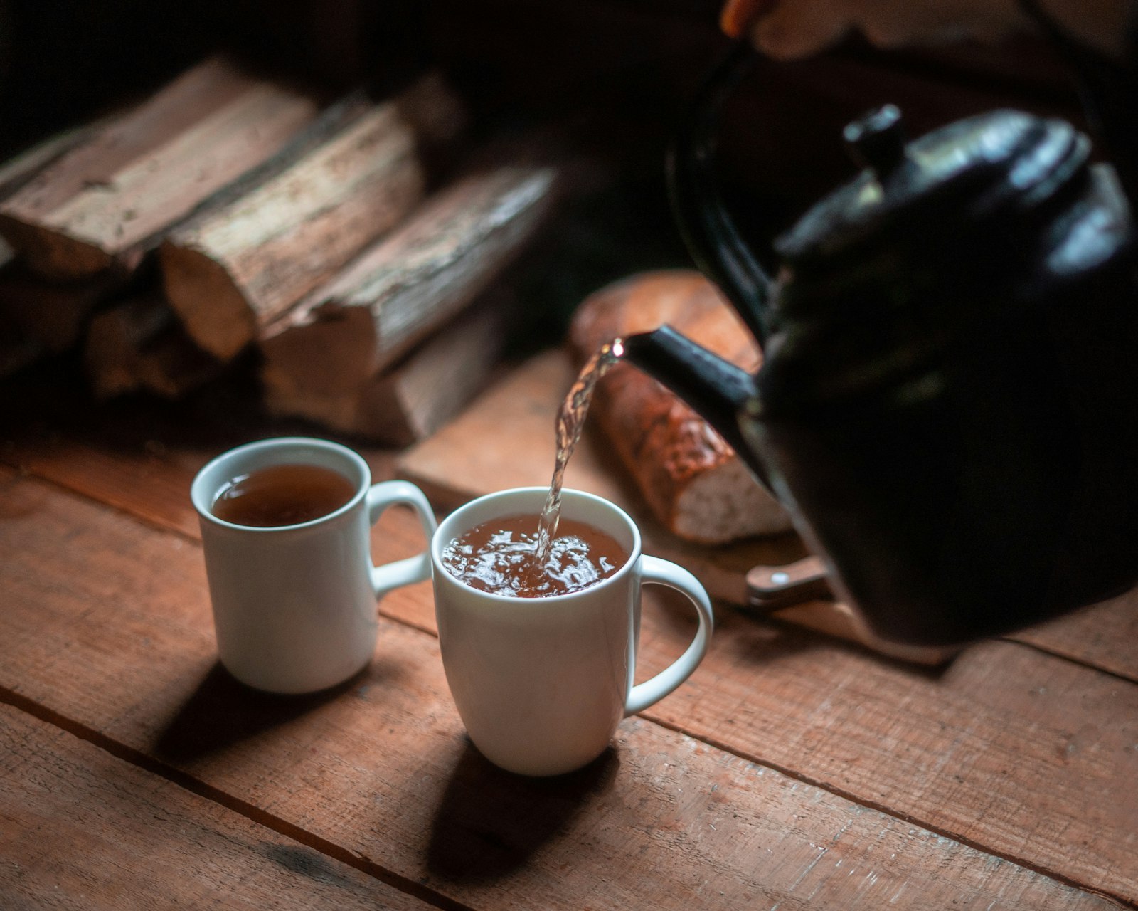 Kettle pouring hot water into two mugs of tea beside firewood in a cabin
