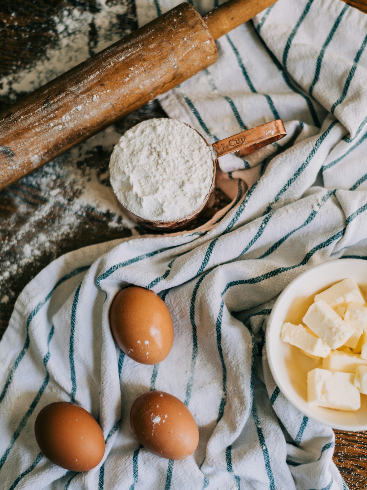 Measuring cup of flour, three eggs and butter on a striped cloth beside a rolling pin