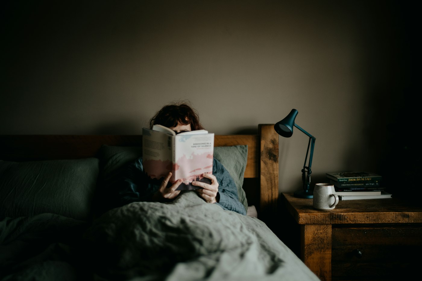 Person reading a book in bed next to a bedside lamp and a stack of books