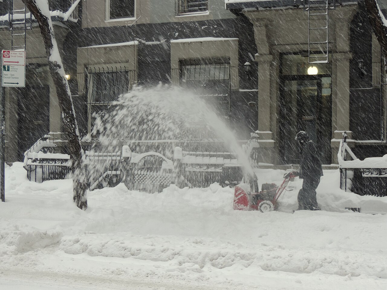 Person using a walk-behind snow blower to clear a sidewalk during a heavy snowfall
