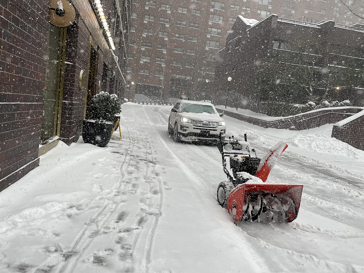 Snow blower parked on a snowy driveway with a car approaching during a snowstorm