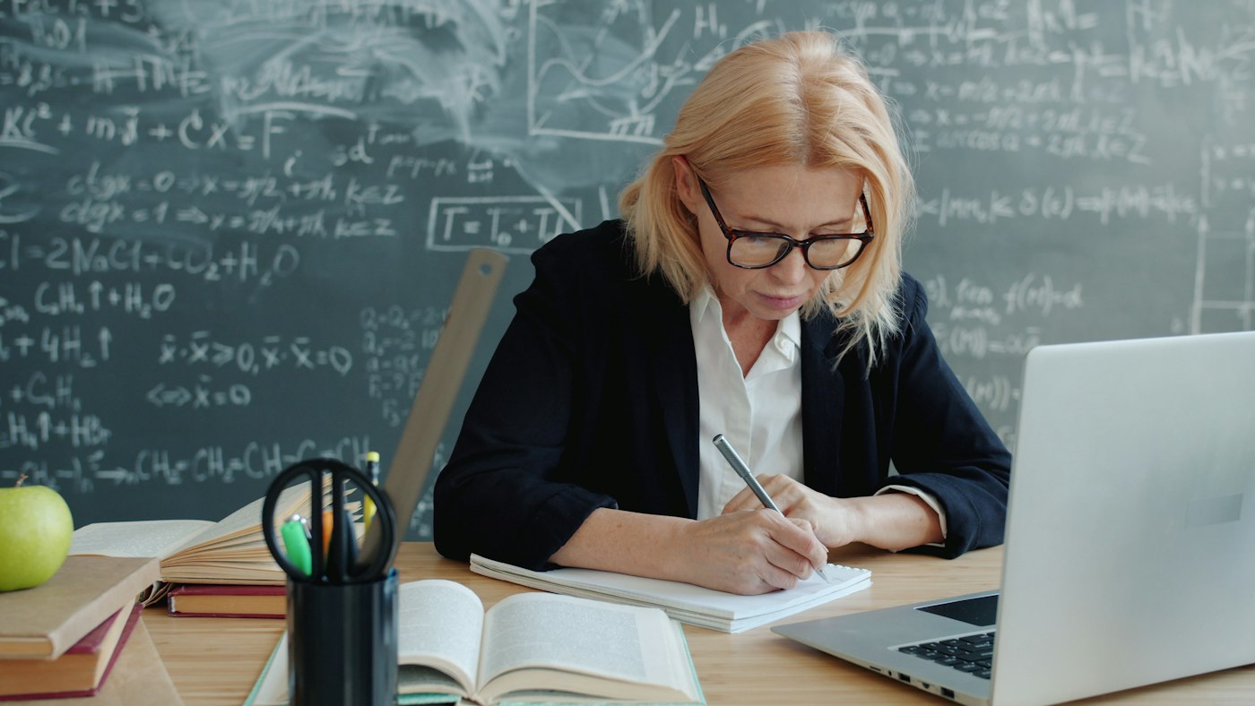 Teacher writing at a desk with books and a laptop in a classroom