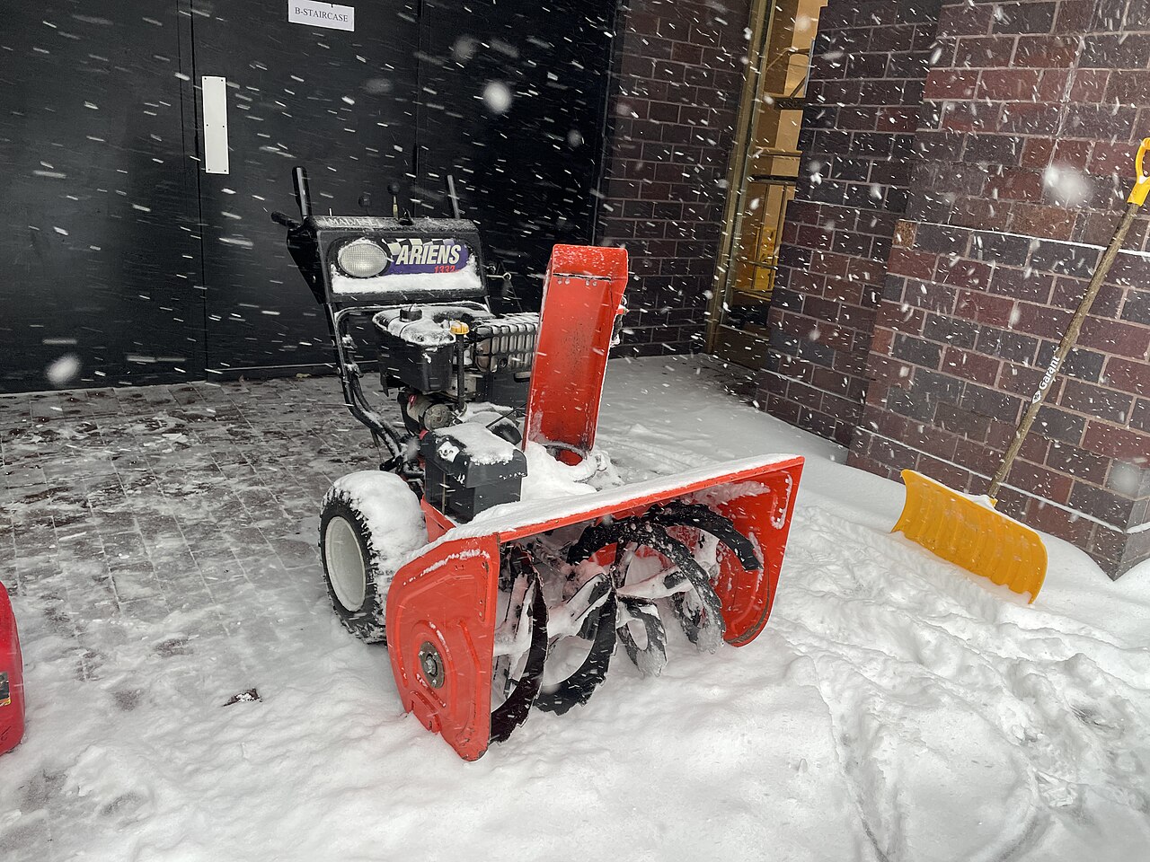 Gas-powered two-stage snow blower with an orange auger housing parked in falling snow